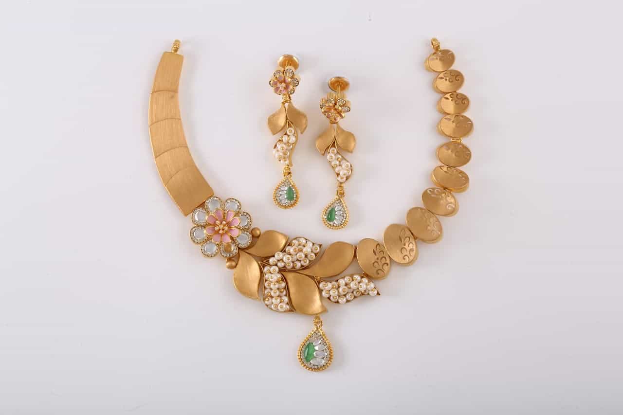 Gold necklace and matching earrings, adorned with pearls, green and white gemstones, and floral designs, displayed on a white background