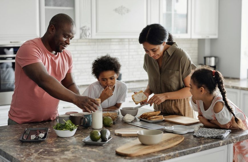 A happy family of four gathered around a kitchen island, preparing breakfast together, with the father reaching for a milk bottle, the mother spreading cream on toast