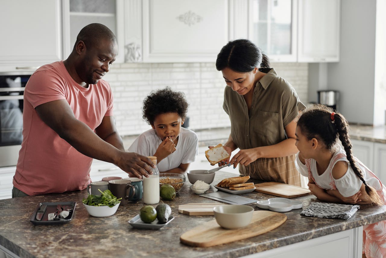 A happy family of four gathered around a kitchen island, preparing breakfast together, with the father reaching for a milk bottle, the mother spreading cream on toast