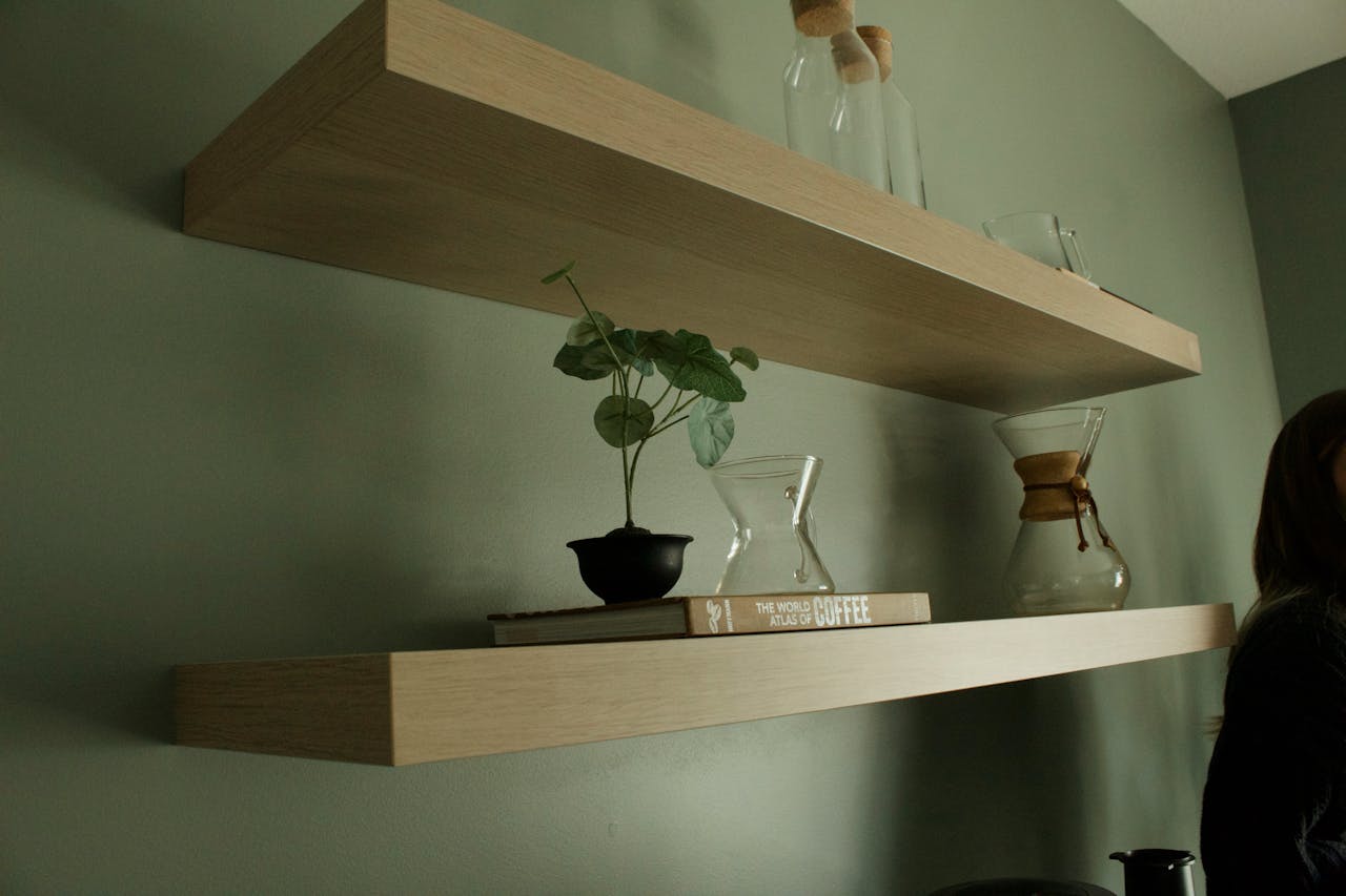 Minimalist wooden floating shelves mounted on a sage green wall, top shelf holds glass bottles with cork stoppers, bottom shelf holds a small potted plant, a coffee dripper