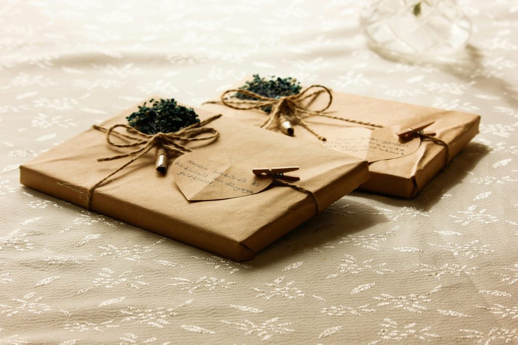 Two neatly wrapped gifts in brown paper, tied with twine bows, decorated with small green flower bundles, heart-shaped tags, and mini clothespins, resting on a lace-patterned white cloth