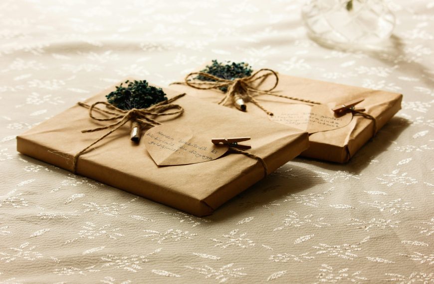 Two neatly wrapped gifts in brown paper, tied with twine bows, decorated with small green flower bundles, heart-shaped tags, and mini clothespins, resting on a lace-patterned white cloth