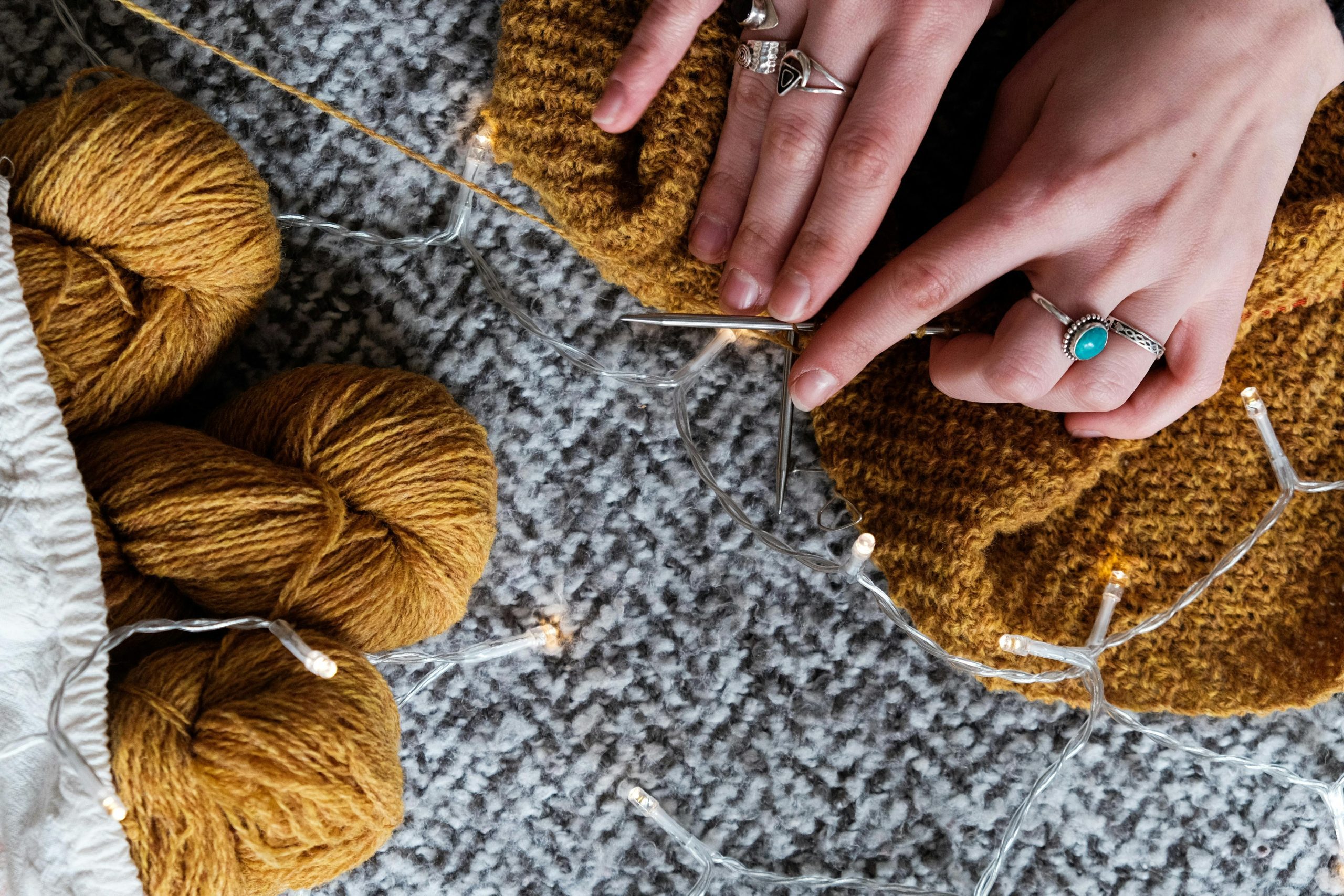 Hands knitting with mustard yellow yarn on a grey textured carpet, metal knitting needles in use, three skeins of matching yarn nearby, string lights spread around the workspace, person wearing silver rings including one with a turquoise stone, cozy and creative setting
