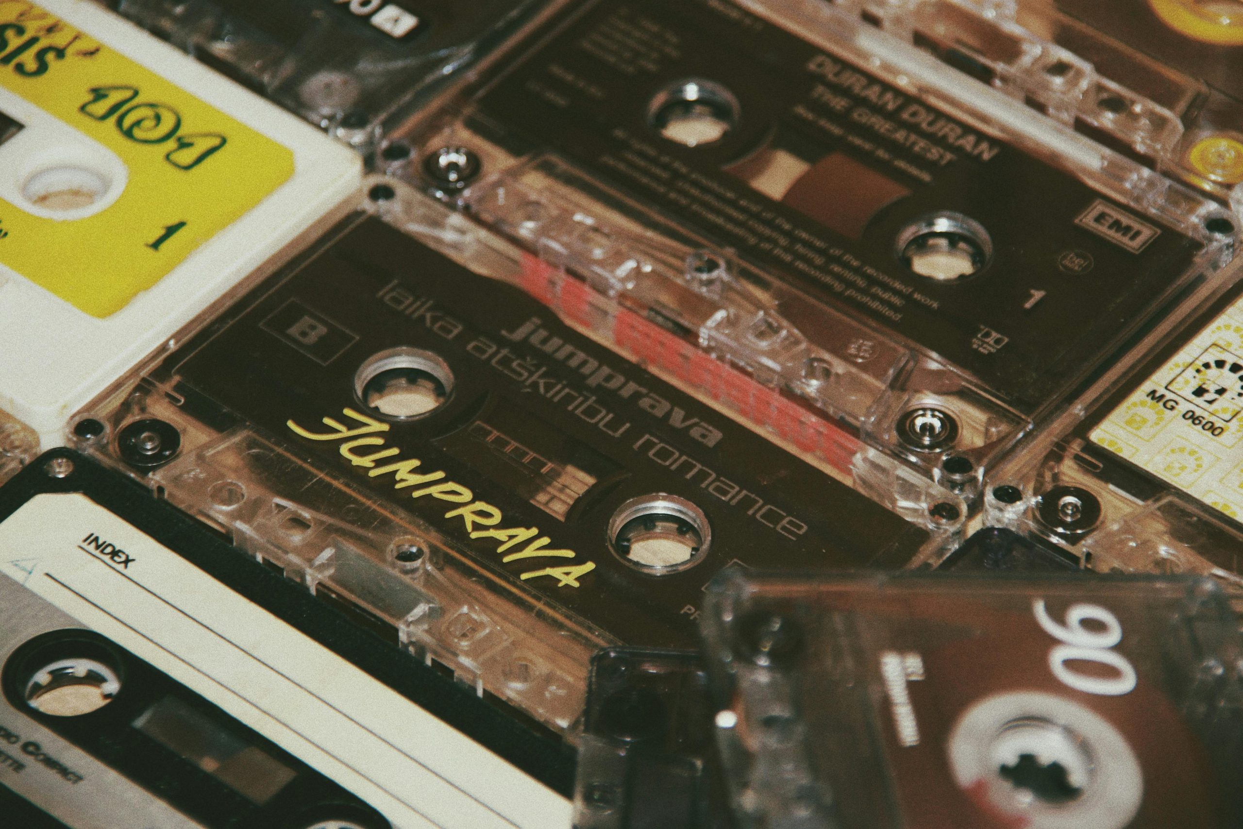 Close-up of a collection of vintage audio cassette tapes, various brands and labels visible including Jumpprava and Duran Duran, stacked in a scattered arrangement, mix of clear, white, and colored plastic casings, retro music theme emphasized by nostalgic design elements