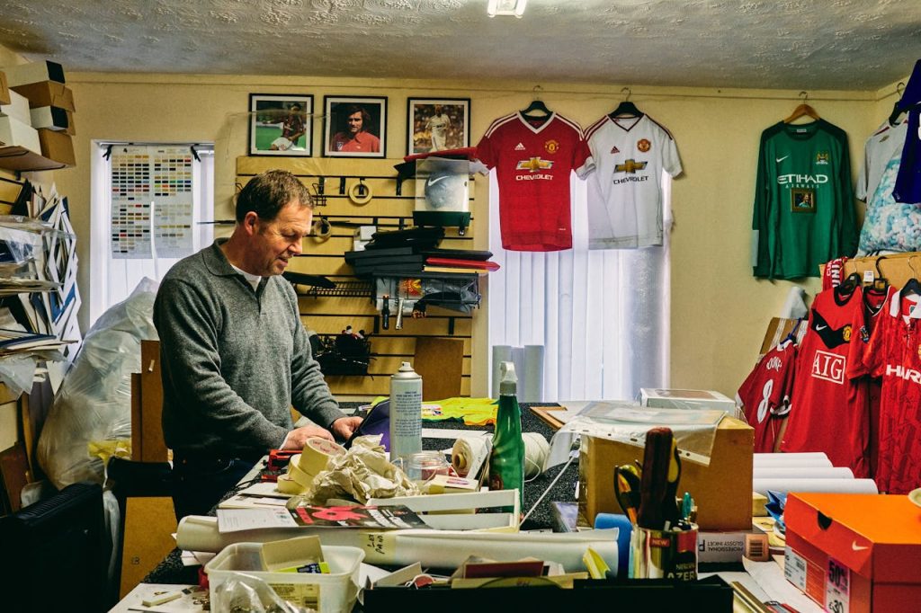 Man working in a cluttered workshop or printing room, surrounded by sports memorabilia, tools, and supplies, with various Manchester United football jerseys and framed photographs hanging on the wall behind him