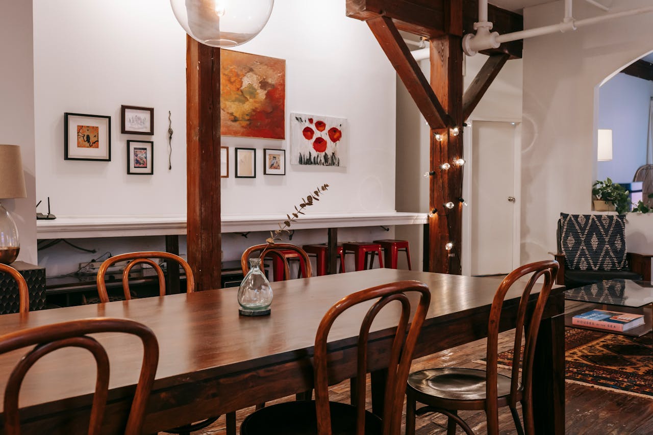 Cozy interior with a long wooden dining table, curved wooden chairs, glass vase with a dried branch centerpiece, white walls with framed artwork, exposed wooden beams with string lights