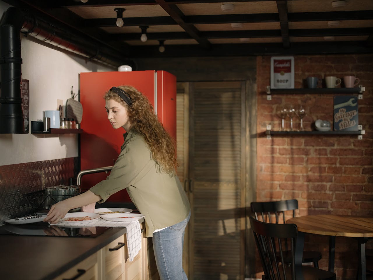 Woman with curly hair setting plates on a kitchen counter, standing near a red refrigerator, in a cozy kitchen with brick walls and wooden decor