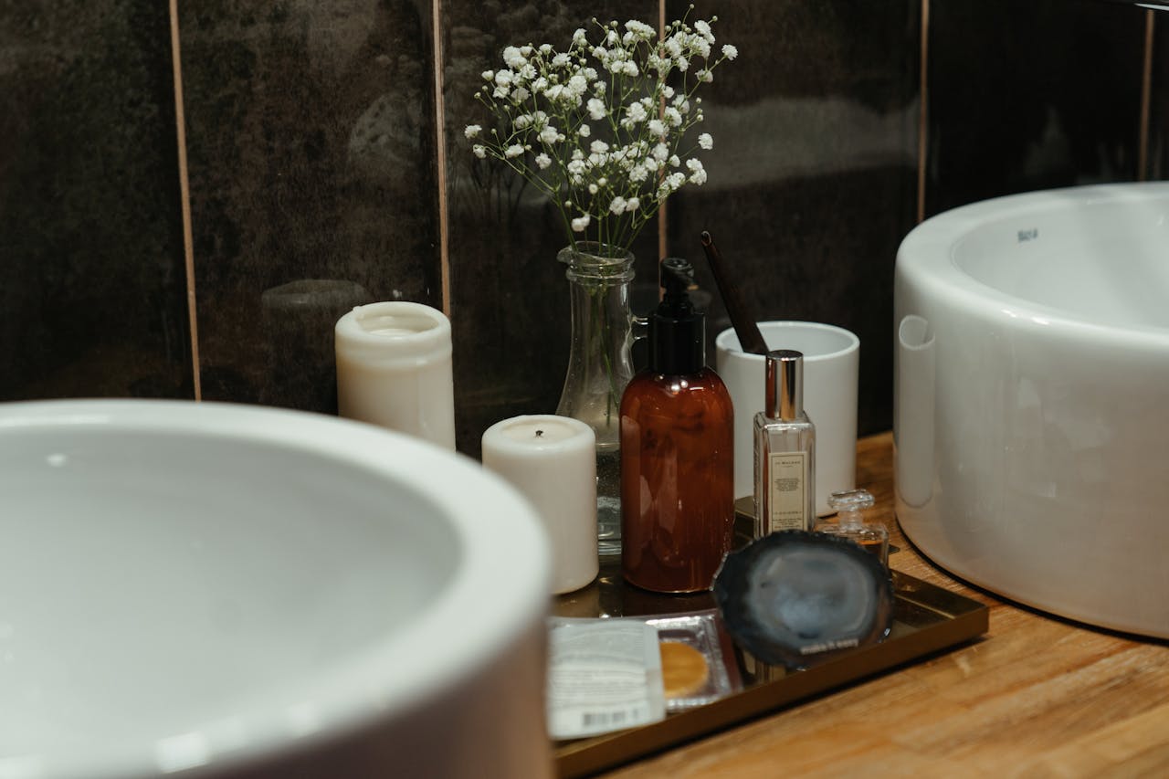 Bathroom countertop with white sinks, candles, amber pump bottle, perfume, skincare products, a toothbrush in a white cup, and a small vase with white baby's breath flowers, all arranged on a tray