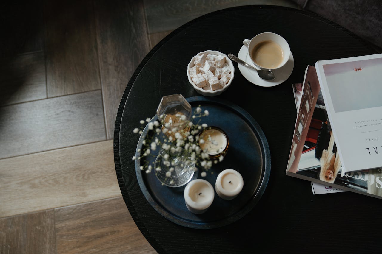 Round black table with a tray holding white candles, a glass vase with baby's breath flowers, a small candle jar, and a drink, beside a cup of coffee with a saucer and spoon, a bowl of powdered sweets, and a stack of fashion and design magazines