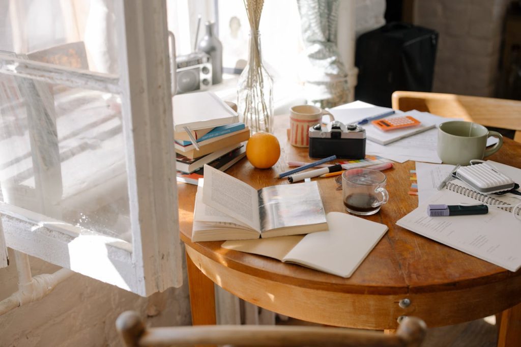 Wooden round table by a sunlit window, covered with open books, pens, a calculator, a coffee cup, a camera, papers, and an orange