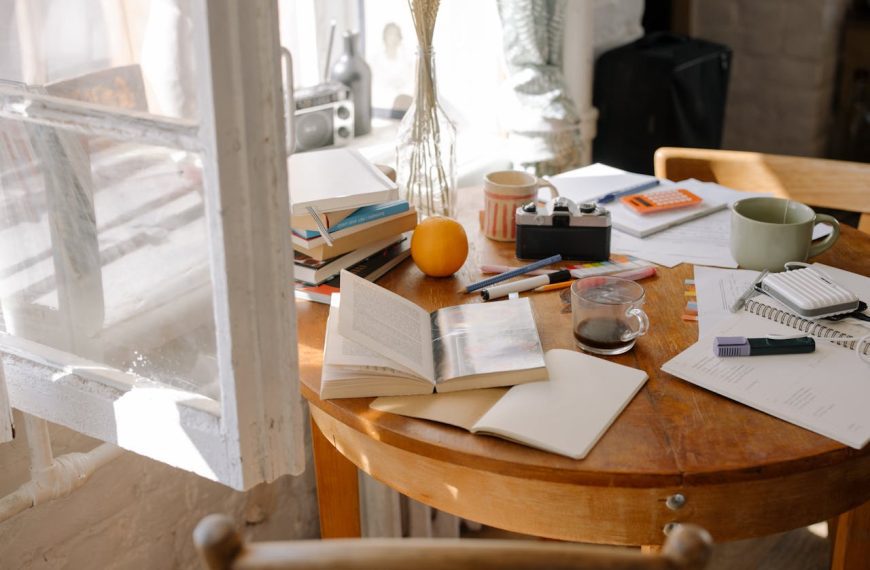 Wooden round table by a sunlit window, covered with open books, pens, a calculator, a coffee cup, a camera, papers, and an orange