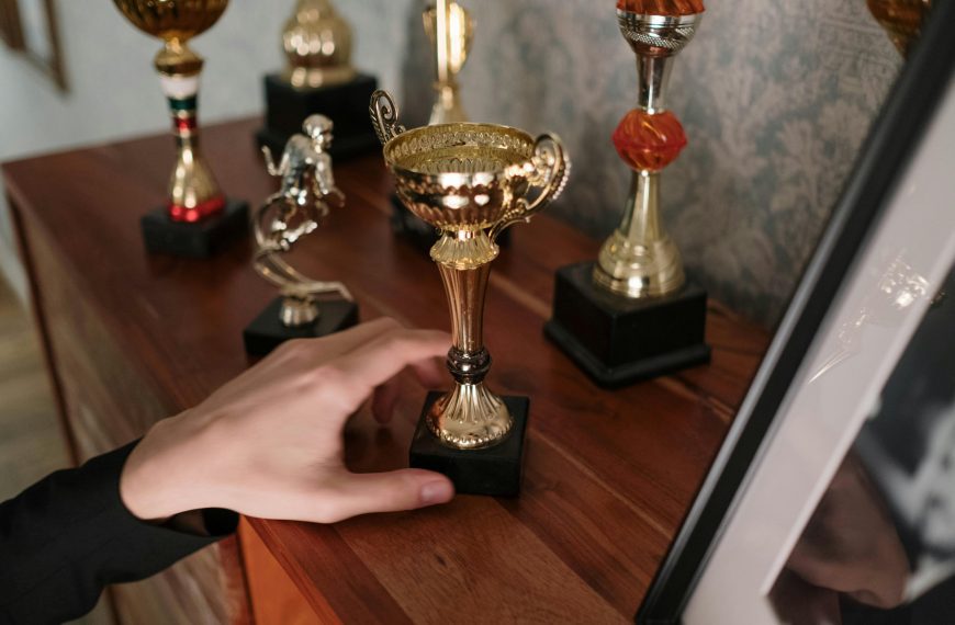 Close-up of a person’s hand reaching for a small gold trophy, several other trophies in different shapes and colors arranged on a wooden shelf, framed photo partially visible, wallpapered background