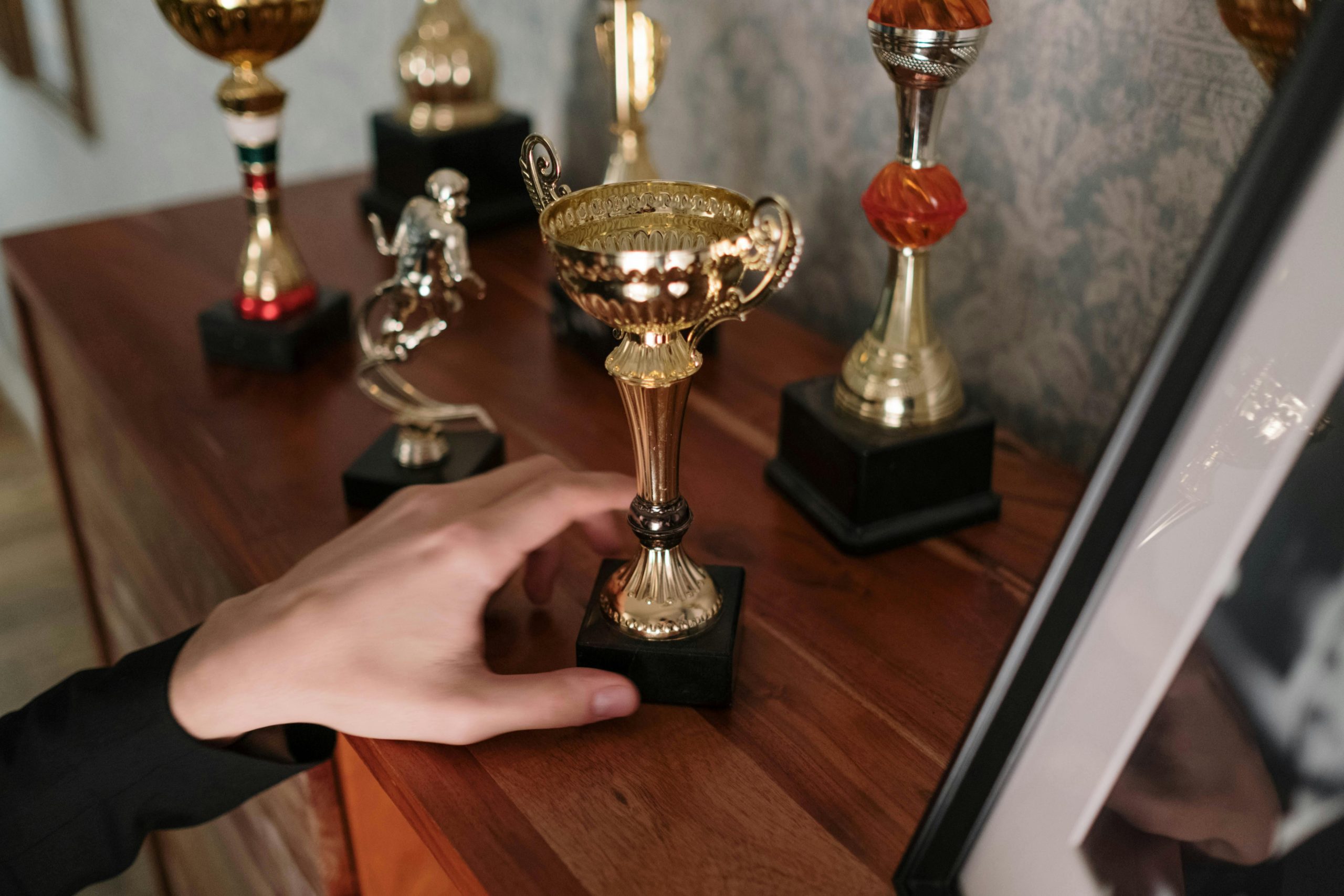 Close-up of a person’s hand reaching for a small gold trophy, several other trophies in different shapes and colors arranged on a wooden shelf, framed photo partially visible, wallpapered background