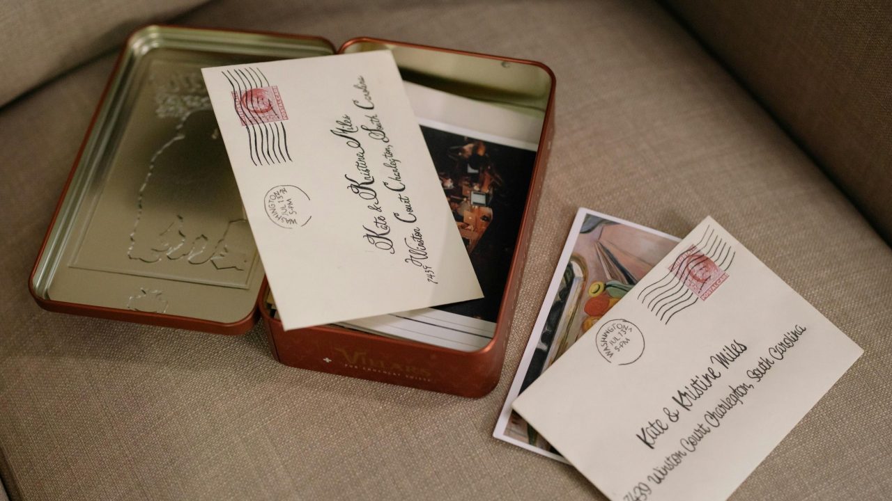 Envelope addressed to Kate and Matthew Miles, placed on a beige couch, one envelope inside a metal tin box along with printed photographs, Washington postmark visible, soft lighting suggests a sentimental or nostalgic moment