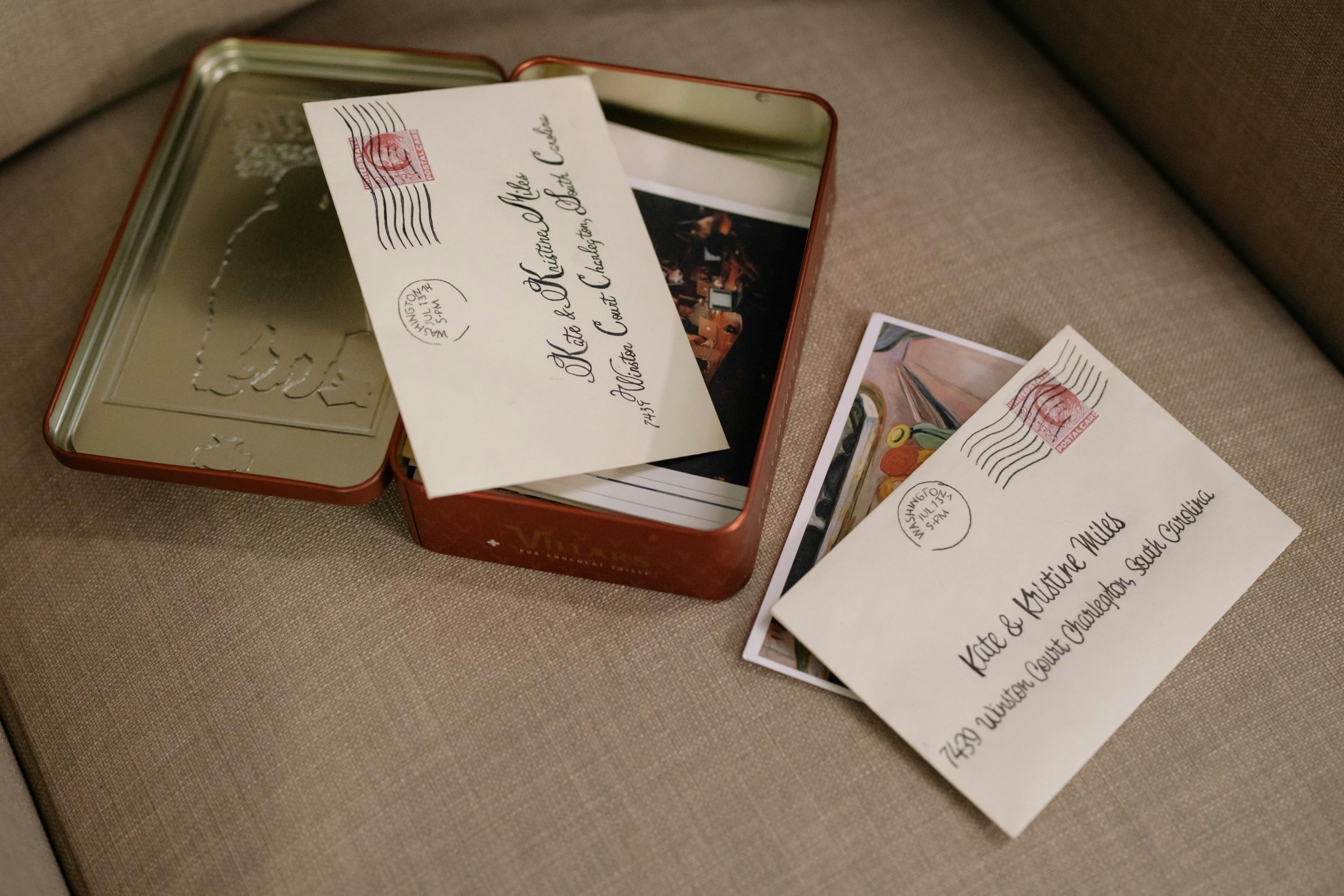 Envelope addressed to Kate and Matthew Miles, placed on a beige couch, one envelope inside a metal tin box along with printed photographs, Washington postmark visible, soft lighting suggests a sentimental or nostalgic moment