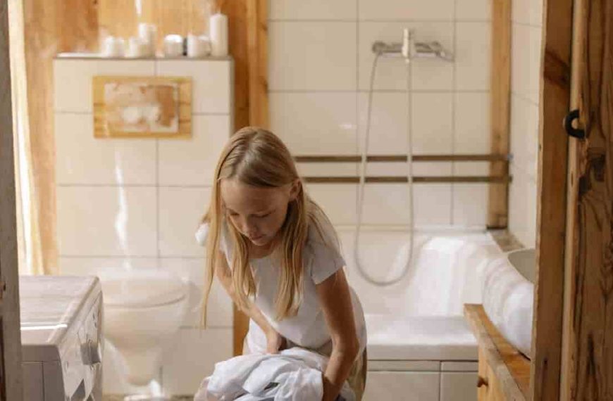 Young girl with long blonde hair, loading clothes into a front-loading washing machine, inside a tiled bathroom with wooden accents and a bathtub in the background
