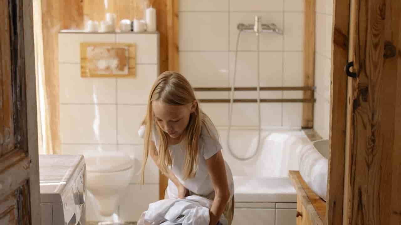 Young girl with long blonde hair, loading clothes into a front-loading washing machine, inside a tiled bathroom with wooden accents and a bathtub in the background