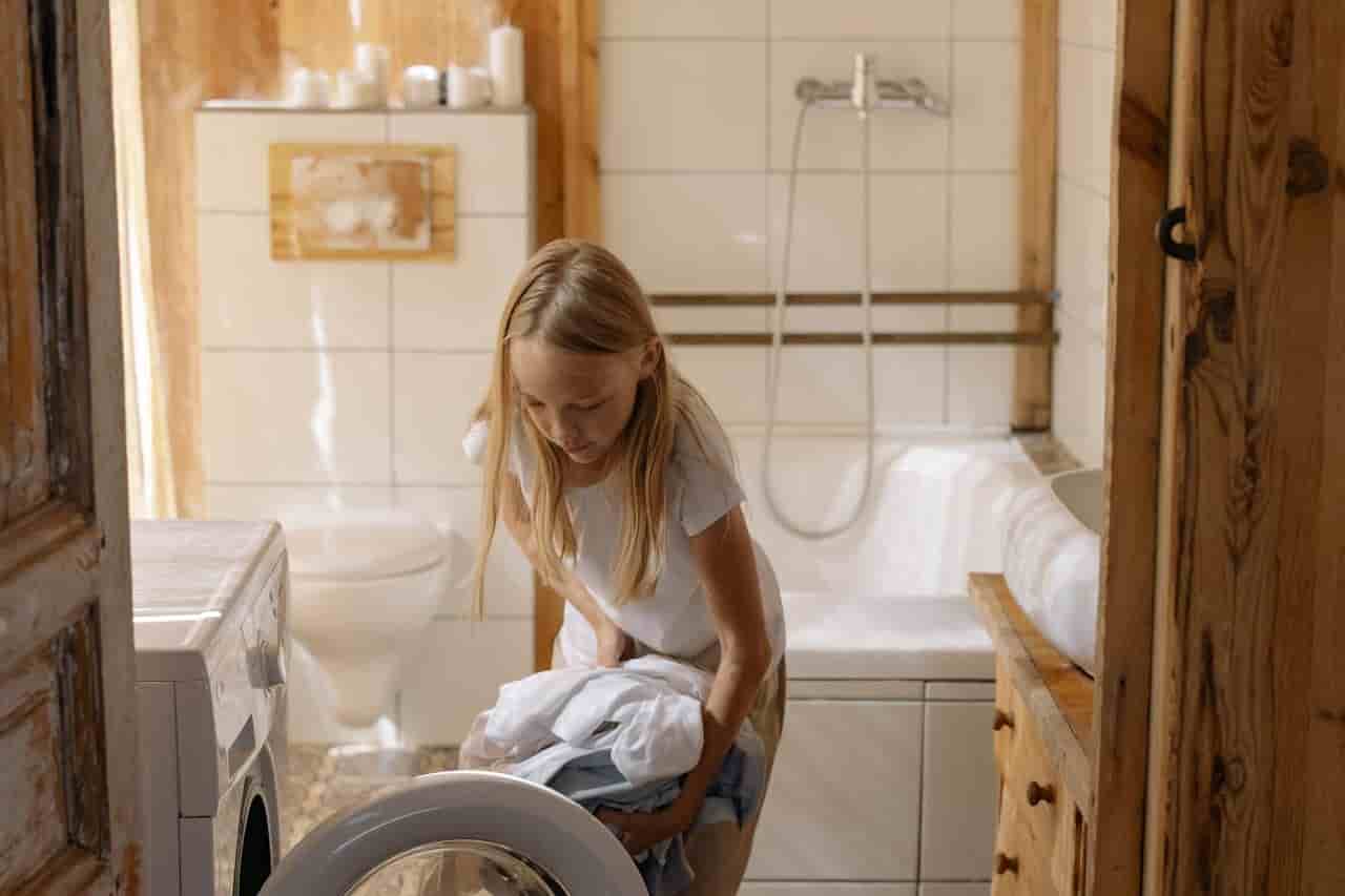 Young girl with long blonde hair, loading clothes into a front-loading washing machine, inside a tiled bathroom with wooden accents and a bathtub in the background