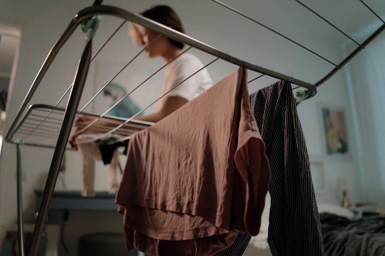 Close-up of clothes drying on a metal indoor drying rack, with a person in the background hanging laundry, soft lighting in a cozy bedroom or living space
