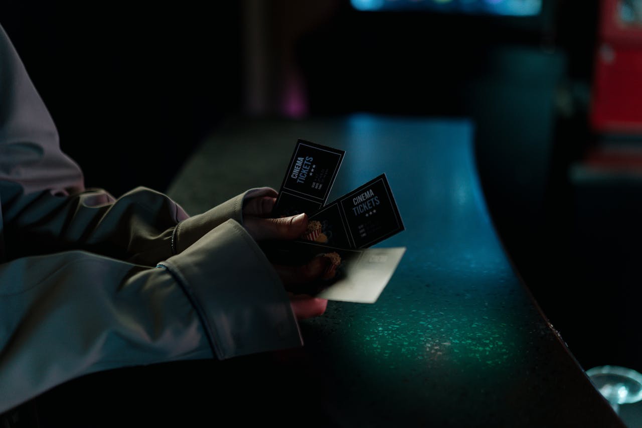 Person wearing a coat holding multiple black cinema tickets, dimly lit setting with green and blue ambient light reflecting off a counter, suggesting a movie theater environment