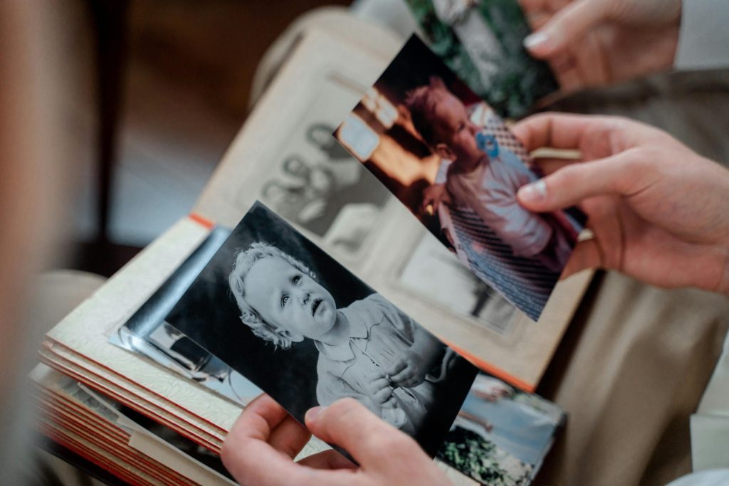 Hands holding old printed baby photos, one in black and white showing a toddler with curly hair, the other in color showing a baby with a pacifier, open photo album on lap with vintage family portraits inside, scene suggests reminiscing or family memory sharing