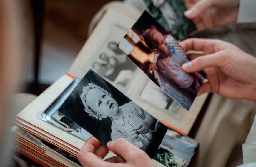 Hands holding old printed baby photos, one in black and white showing a toddler with curly hair, the other in color showing a baby with a pacifier, open photo album on lap with vintage family portraits inside, scene suggests reminiscing or family memory sharing