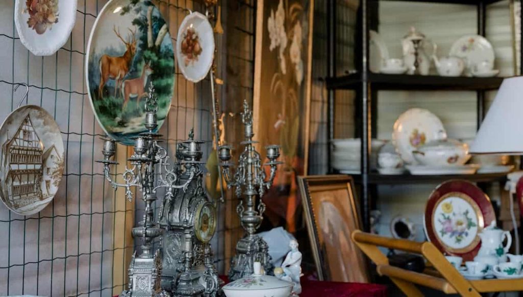 Antique shop display, hanging decorative plates with animal and building scenes, ornate silver candelabras and clock, vintage porcelain tea sets and bowls on shelves, classic artwork in wooden frames, warm ambient lighting
