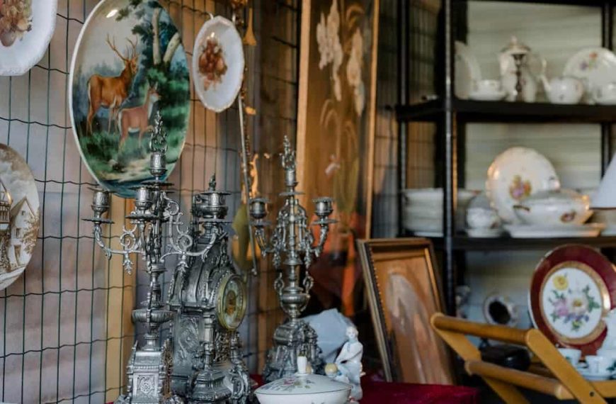 Antique shop display, hanging decorative plates with animal and building scenes, ornate silver candelabras and clock, vintage porcelain tea sets and bowls on shelves, classic artwork in wooden frames, warm ambient lighting