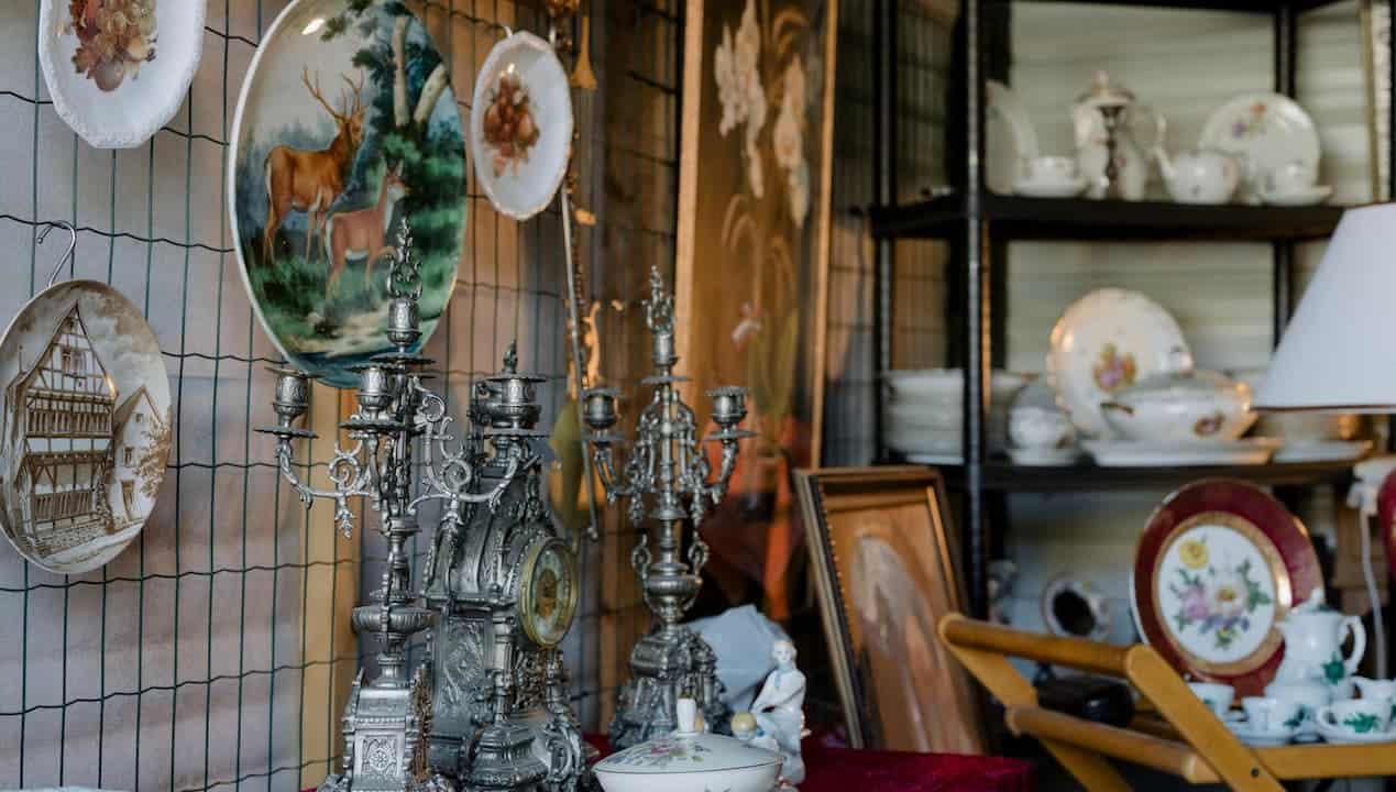 Antique shop display, hanging decorative plates with animal and building scenes, ornate silver candelabras and clock, vintage porcelain tea sets and bowls on shelves, classic artwork in wooden frames, warm ambient lighting