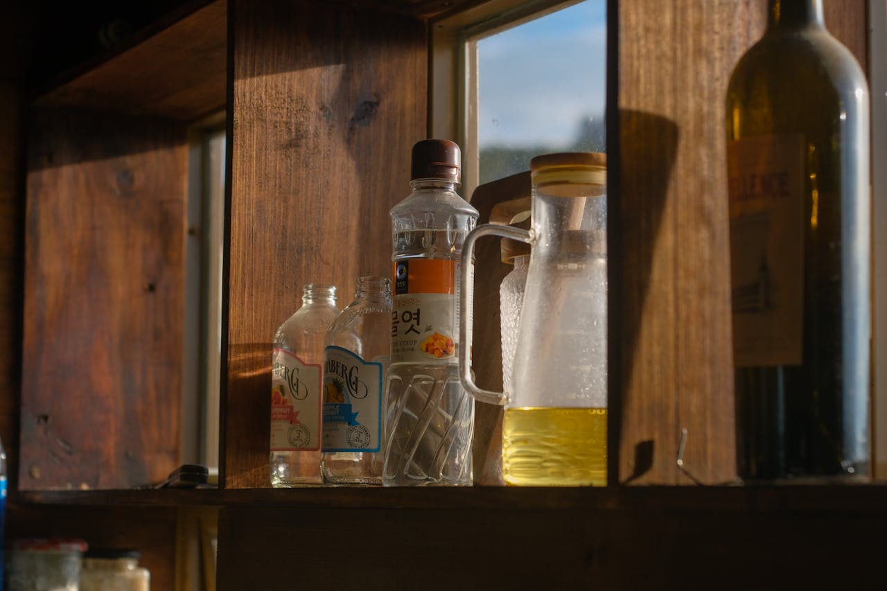 Wooden kitchen shelf by a window, holding glass bottles and jars including clear drinks, a bottle of oil, and a jug with yellow liquid, lit warmly by natural sunlight