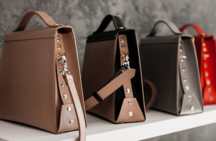 Row of structured leather handbags in various colors including taupe, brown, black, grey, and red, displayed on a white shelf, against a textured grey wall, each bag featuring a top handle and side strap detailing