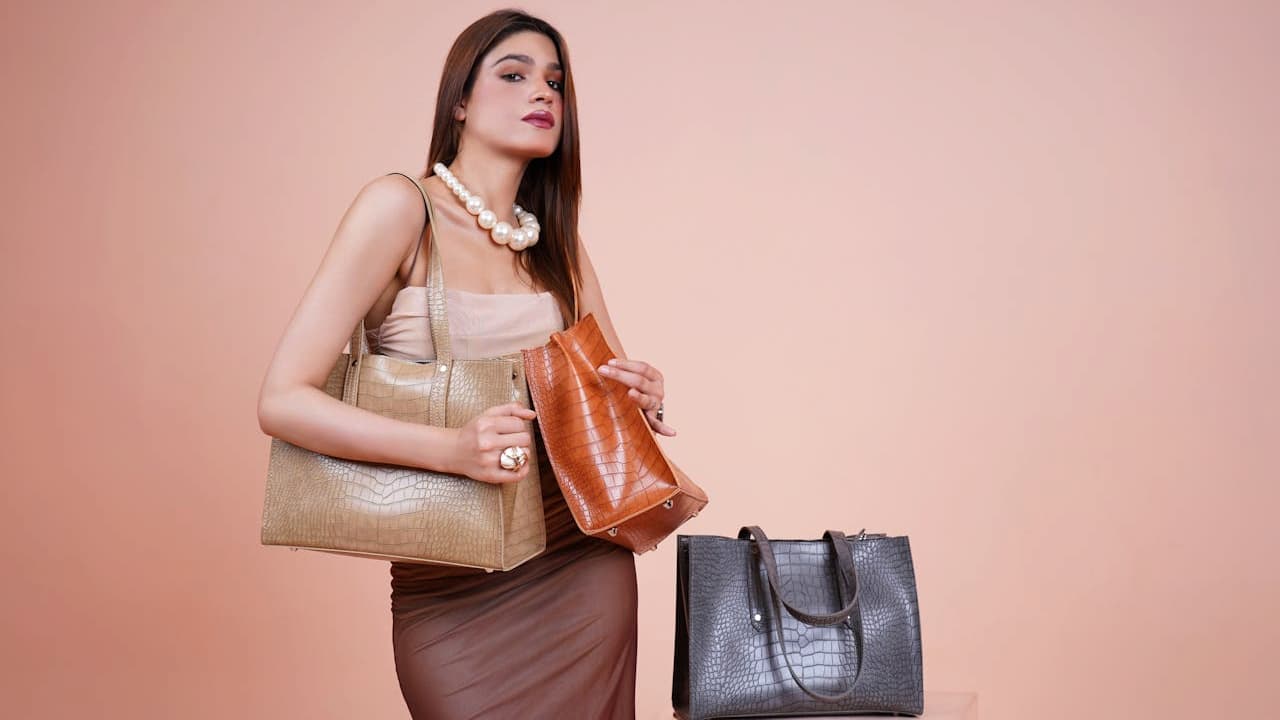Woman posing against a peach background, wearing a beige top and brown skirt, holding a tan handbag in her left hand and an orange handbag in her right, with a black handbag and a gray handbag placed in front of her