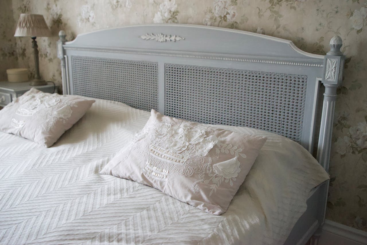 Light blue vintage-style bed with carved wooden headboard and woven rattan panels, floral-embroidered beige pillows, white chevron-patterned bedspread