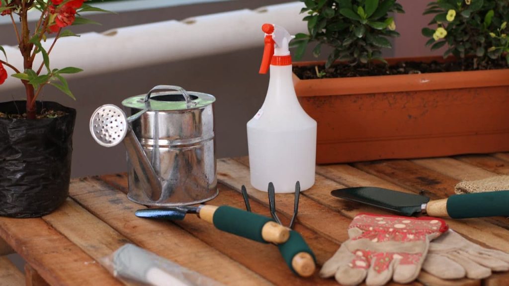 Gardening tools on wooden surface, hand trowel with green handle, garden fork, spray bottle, metal sifter, seed packets, potted plant, rectangular planter in background