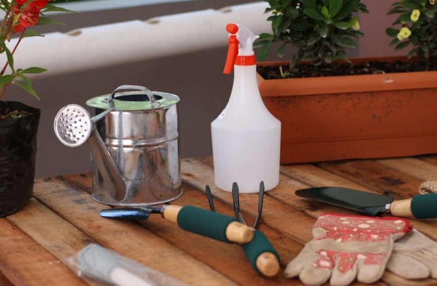 Gardening tools on wooden surface, hand trowel with green handle, garden fork, spray bottle, metal sifter, seed packets, potted plant, rectangular planter in background