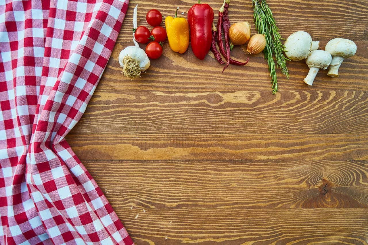Red and white checkered cloth, fresh garlic, cherry tomatoes, yellow and red bell peppers, dried red chilies, onions, rosemary sprigs, white mushrooms, wooden table background