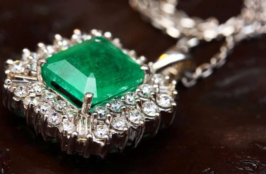 Close-up of a silver necklace with a large square-cut emerald gemstone surrounded by small round diamonds, resting on a dark textured surface