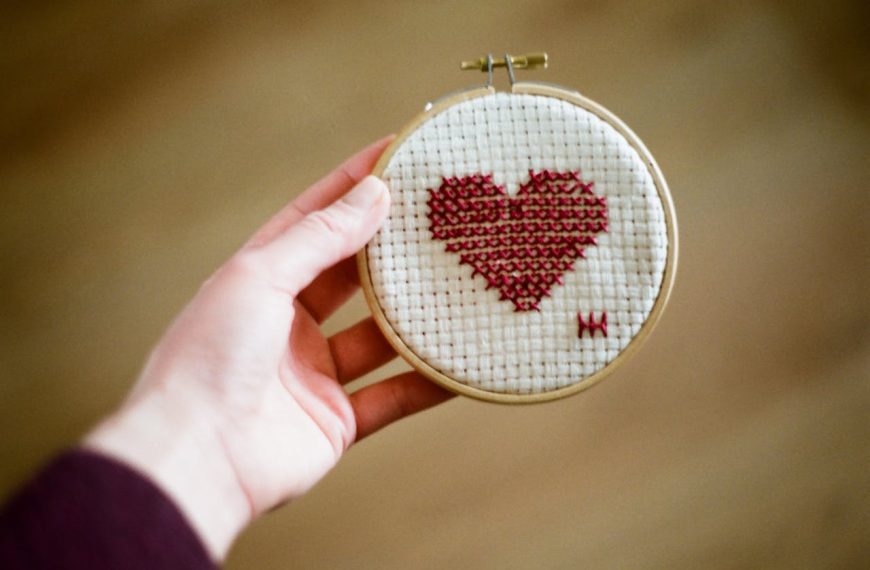 Hand holding a small embroidery hoop with a cross-stitched red heart on white fabric, minimal background