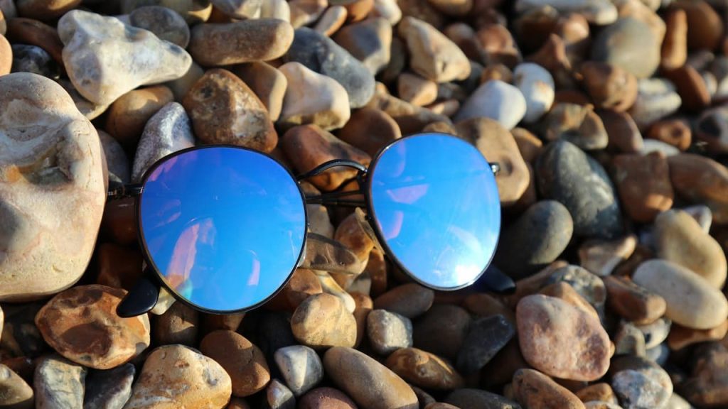 Blue mirrored sunglasses, lying on a bed of multicolored pebbles, reflecting the sky and clouds, outdoor setting, casual and relaxed atmosphere
