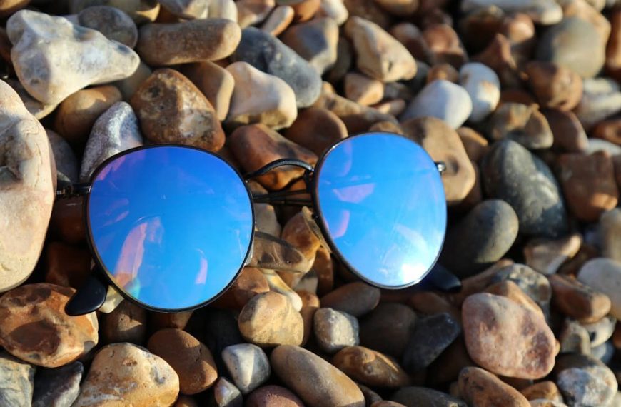 Blue mirrored sunglasses, lying on a bed of multicolored pebbles, reflecting the sky and clouds, outdoor setting, casual and relaxed atmosphere