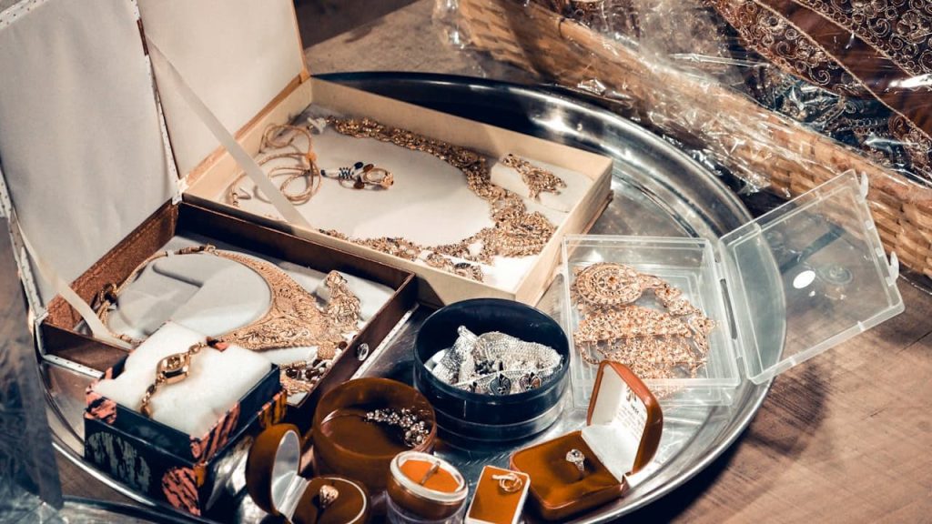 Assorted vintage jewelry on a silver tray, including gold necklaces, rings in velvet boxes, beaded pieces, and ornate sets in open display boxes, placed on a wooden surface with a woven basket in the background
