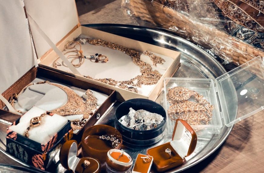 Assorted vintage jewelry on a silver tray, including gold necklaces, rings in velvet boxes, beaded pieces, and ornate sets in open display boxes, placed on a wooden surface with a woven basket in the background