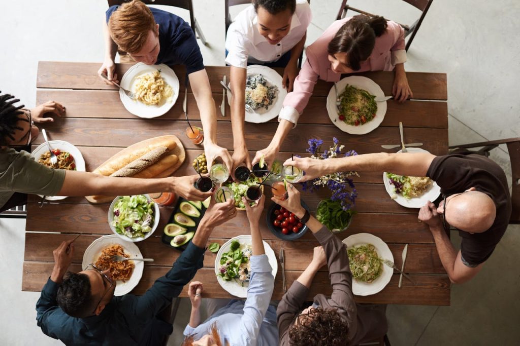 Group of people sitting around a wooden dining table, raising their glasses for a toast, surrounded by plates of pasta, salad, bread, and fresh vegetables