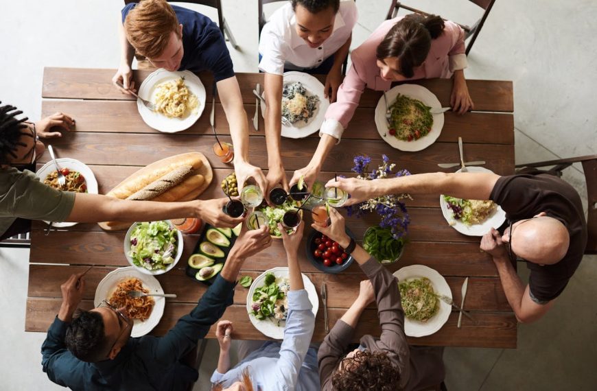 Group of people sitting around a wooden dining table, raising their glasses for a toast, surrounded by plates of pasta, salad, bread, and fresh vegetables