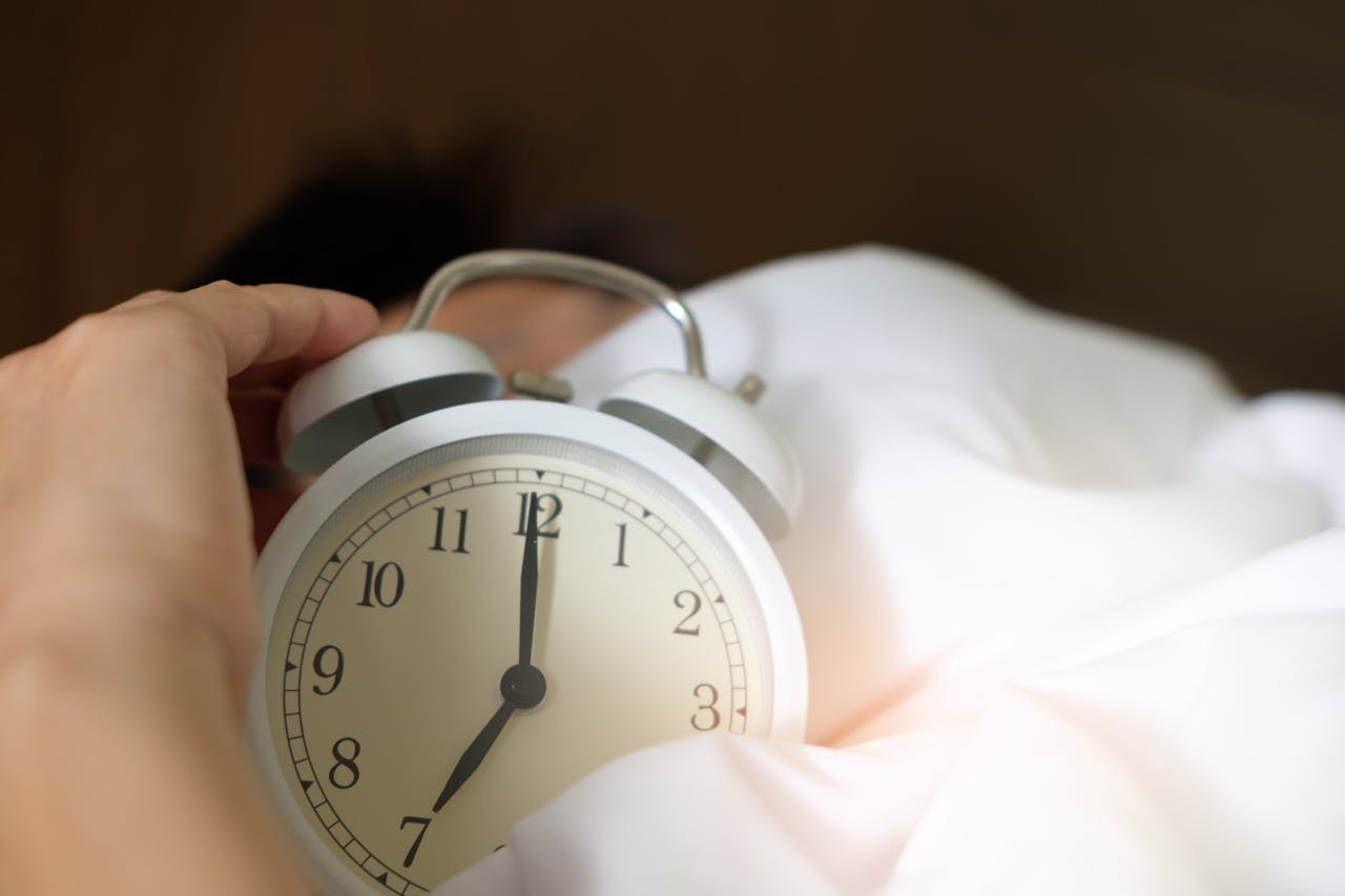 Close-up of a white analog alarm clock showing 7:00, held by a hand over white bed sheets, with a partially visible person lying in bed