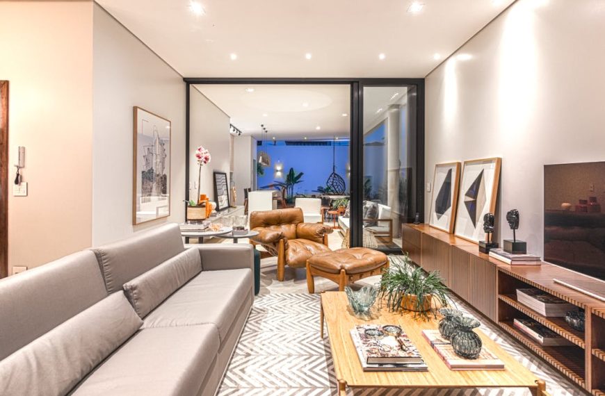 Modern living room interior, gray sofa, leather armchairs, wooden coffee table with decor, TV console with artwork, patterned rug, indoor plants, open layout leading to a dining area