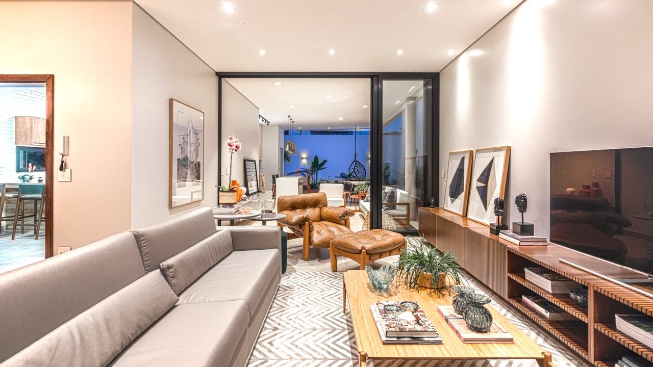 Modern living room interior, gray sofa, leather armchairs, wooden coffee table with decor, TV console with artwork, patterned rug, indoor plants, open layout leading to a dining area