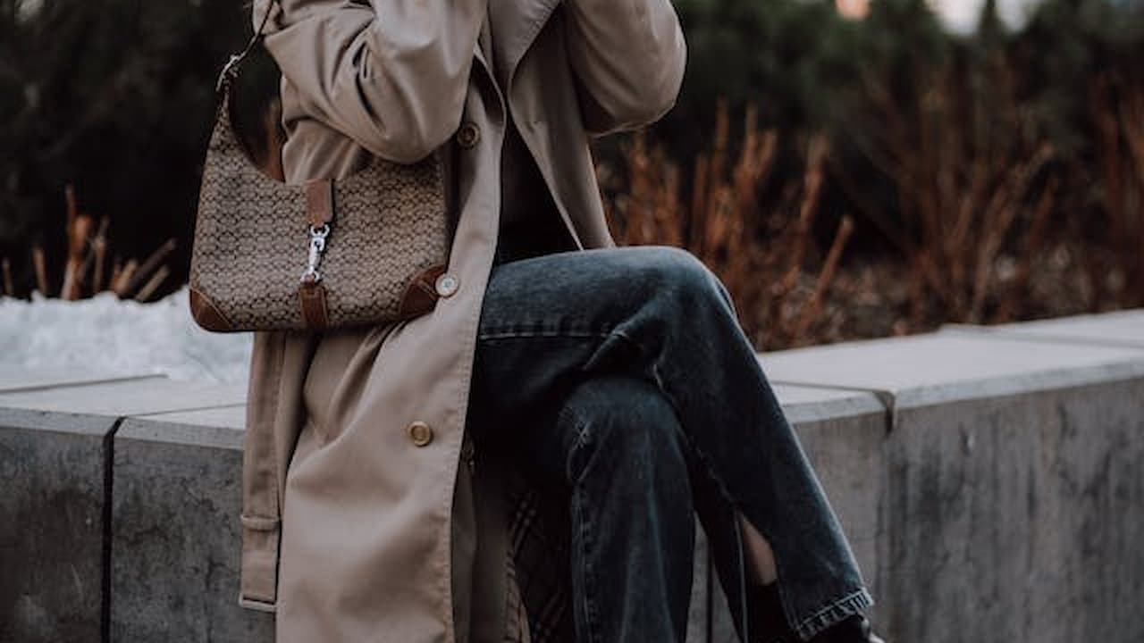 Woman sitting on a concrete ledge, wearing a beige trench coat and dark jeans, carrying a patterned shoulder bag, holding a cup, surrounded by dried plants and greenery in an outdoor urban setting