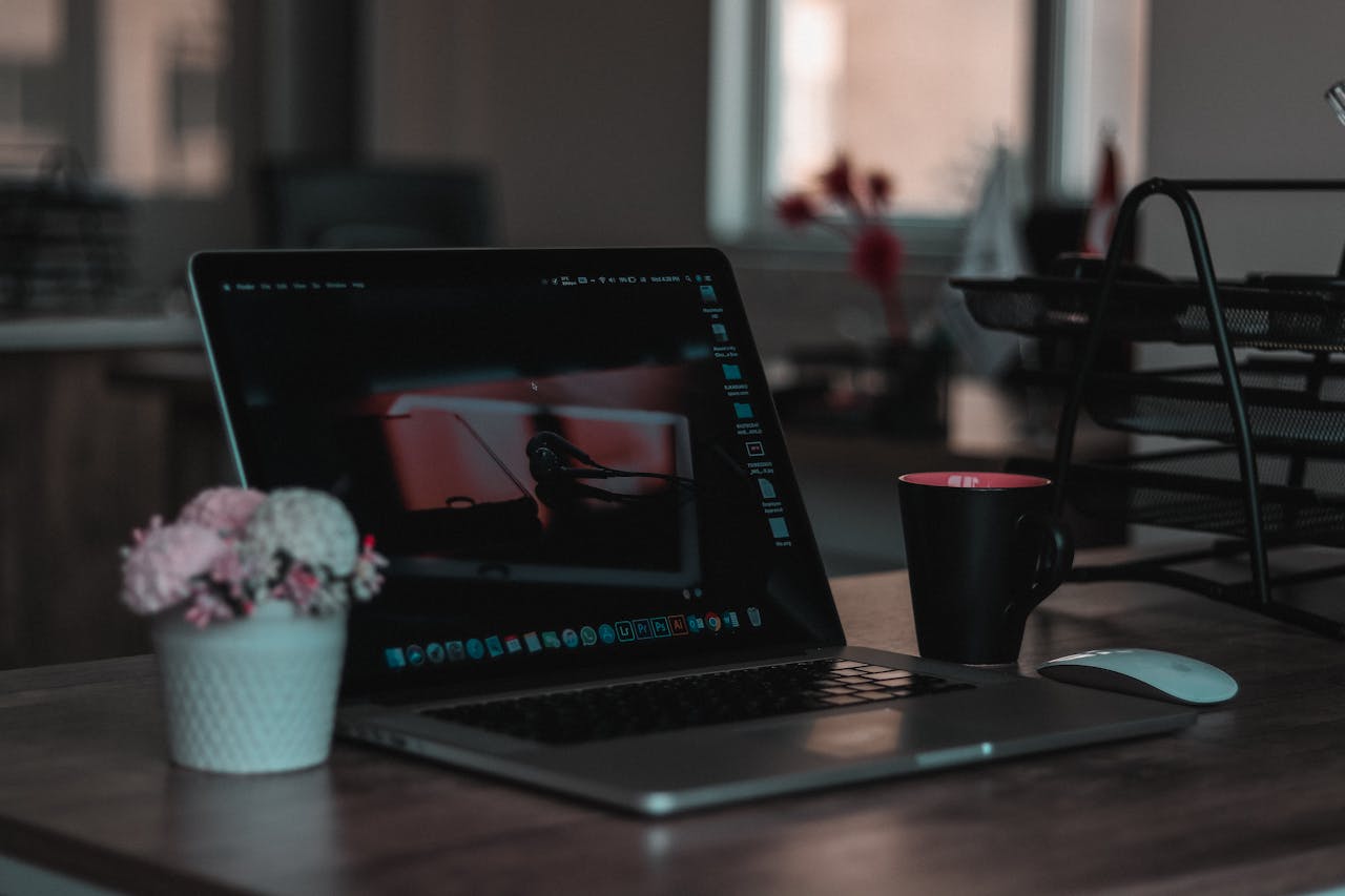 Open laptop on a wooden desk, dark-themed screen with earphones wallpaper, black mug with pink interior, white wireless mouse, potted flowers, and black metal file trays in a softly lit office