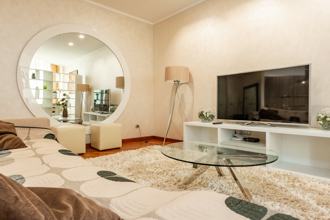 Modern living room, round wall mirror reflecting a cozy space, TV on a white stand, floor lamp, glass coffee table on a fluffy rug, beige walls, patterned couch, small stools in front of mirror